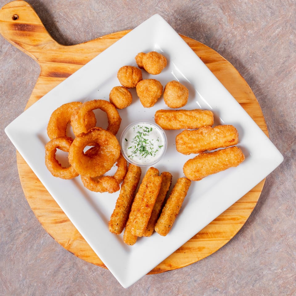 A white square plate of Manhattan Pizza appetizers, including onion rings, mozzarella sticks, and fried mushrooms with ranch.