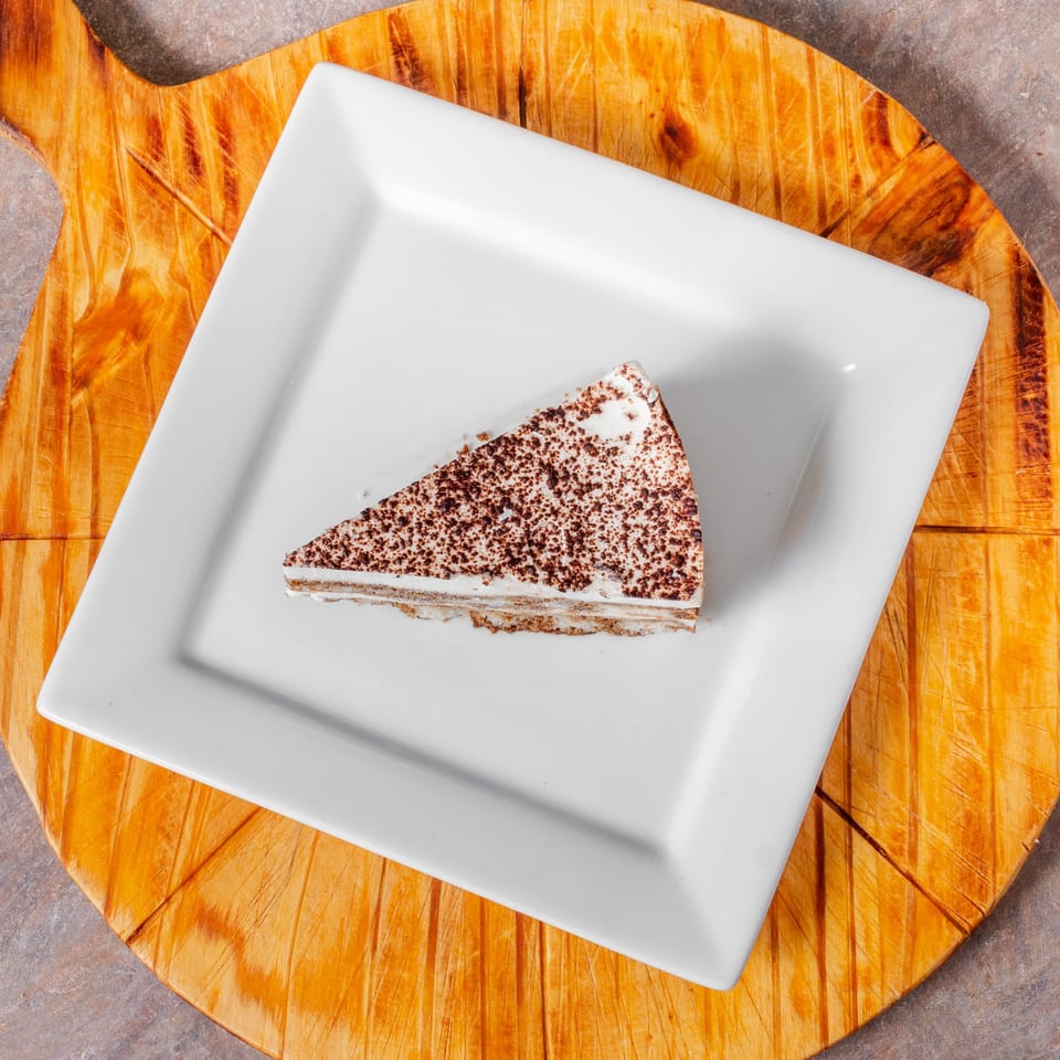A slice of tiramisu cake with chocolate dusting on a white square plate, on a wooden board.