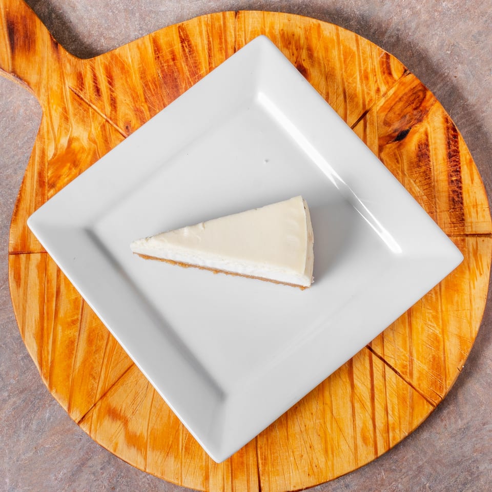 A slice of Manhattan Pizza's classic cheesecake on a white square plate, atop a wooden board.