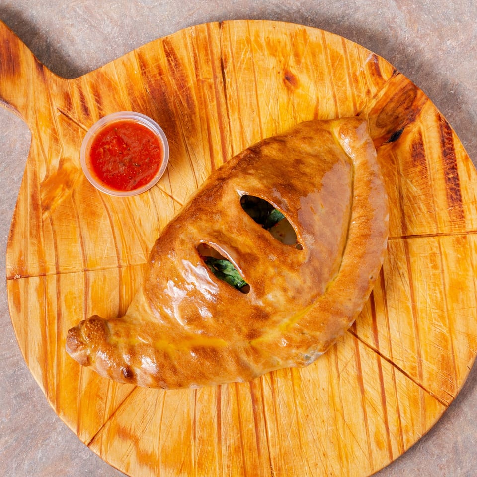 Manhattan Pizza calzone with visible spinach and a side of marinara sauce on a wooden board.